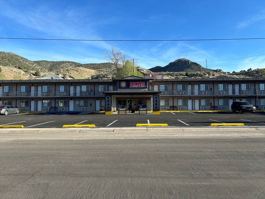 Un motel con una estructura de dos pisos, un estacionamiento al frente y montañas al fondo.