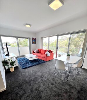 Una sala de estar moderna con un sofá rojo, una mesa de centro blanca y un tapete azul. Hay una zona de comedor con una mesa y sillas blancas. La habitación tiene ventanas amplias y una lámpara de techo.