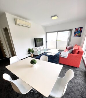 Una sala de estar moderna con una mesa blanca, sillas blancas, un sofá rojo, un televisor de pantalla plana y una pequeña planta.