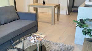 A modern living room with a grey sofa, a glass coffee table with magazines, a wooden dining table, and a kitchen area in the background. - Splendid 2bd 1bth 1csp Apt - Superb CBD Location (Reid)