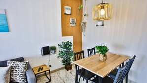 A modern living room with a wooden dining table, black chairs, a grey sofa, a patterned throw pillow, a small wooden side table, a remote control, a television, and a hanging light fixture. - Condo 3 min. from beach with A/C and balcony (Loulé)