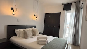 A neatly made bed with pillows and a blanket, a dark headboard, a wall-mounted light fixture, a wardrobe, and a window with curtains. - Condo 3 min. from beach with A/C and balcony (Loulé)