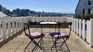 Ein Sitzbereich im Freien mit zwei Stühlen und einem Tisch, einem weißen Zaun und einer Aussicht auf Gebäude und Boote in der Ferne.
