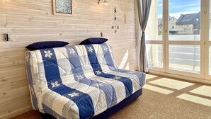 A cozy room with a wooden wall, a large window with curtains, a quilted sofa, and a small shelf with decorative items.