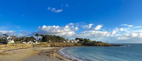 A coastal beach with a clear sky, rocky shoreline, and houses in the background.