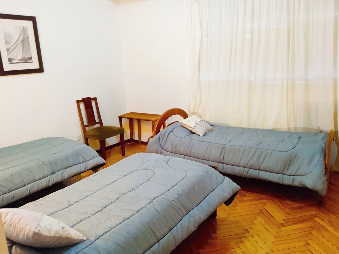 Une chambre avec deux lits simples, une chaise en bois, une petite table et un tableau encadré au mur.
