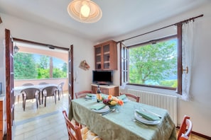 Un coin repas avec une table dressée pour quatre personnes, un téléviseur posé sur un meuble et une vue sur la verdure à l’extérieur.