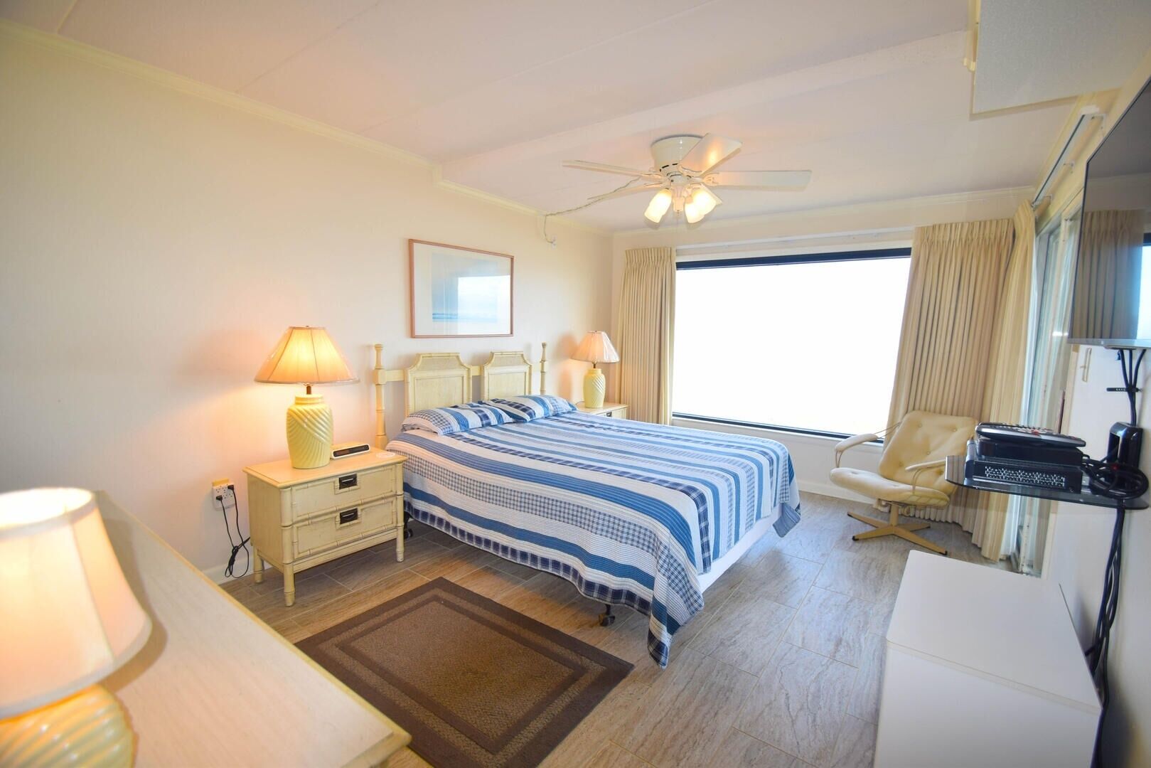 A hotel room with a bed, two bedside tables with lamps, a chair, a desk with a computer, a ceiling fan, and a window with curtains.