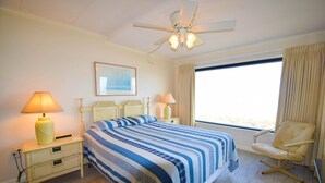 Ein Hotelzimmer mit einem Bett, Nachttischlampen, einem Sessel, einem Deckenventilator, einem Fenster mit Vorhängen und einem gerahmten Bild an der Wand.