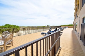 A wooden deck with lounge chairs and a small table, overlooking a beach and a fence.