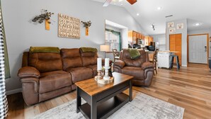 A spacious living room with a brown leather sofa, a wooden coffee table, and a ceiling fan. A kitchen is visible in the background. - Next to Silver Dollar City - Lakeside Condo (Branson)