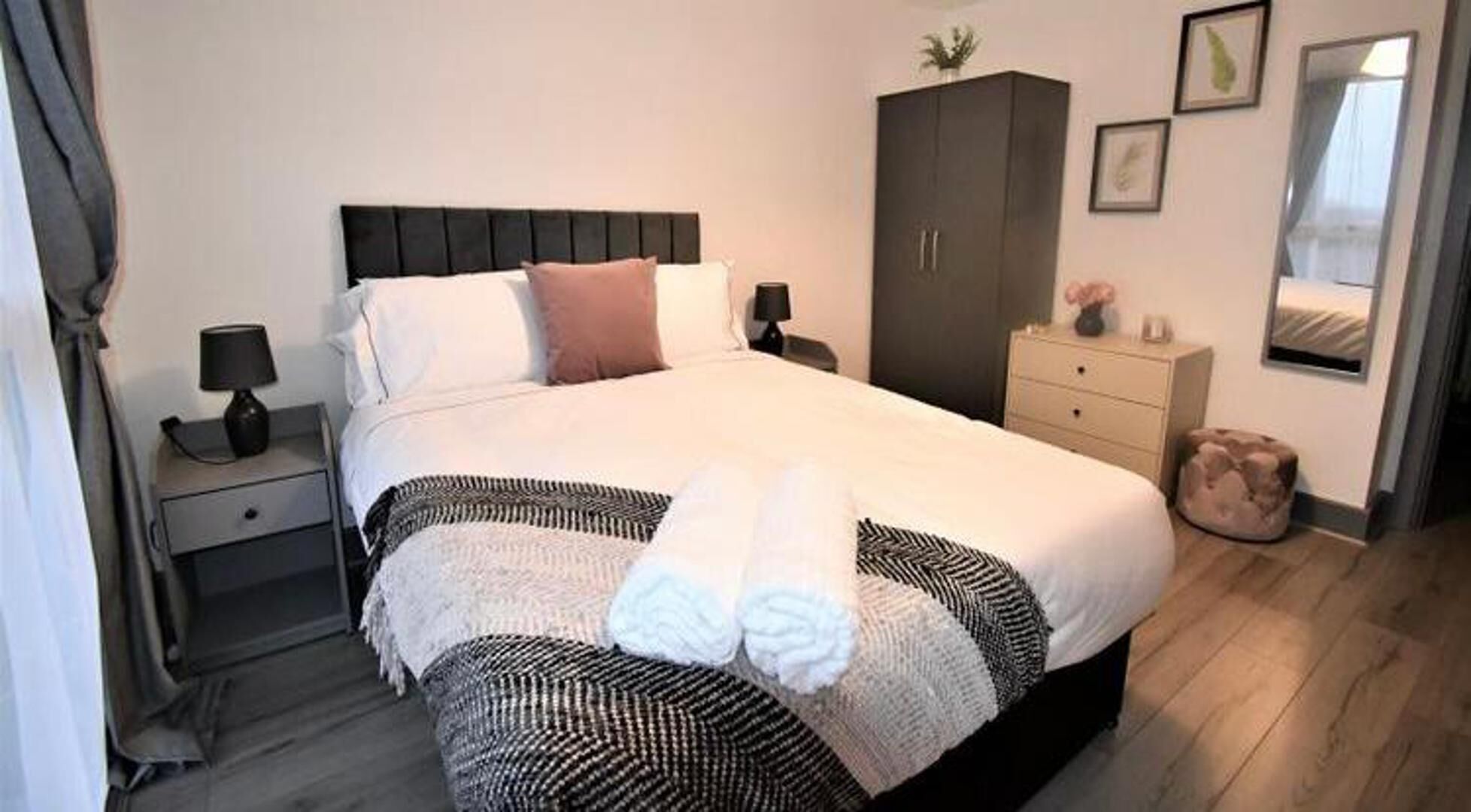 Un letto rifatto con una coperta a righe bianche e nere, un cuscino bianco e un cuscino decorativo marrone.  Un comodino con una lampada e un tavolino con un vaso sono visibili. Sullo sfondo, un armadio e uno specchio.