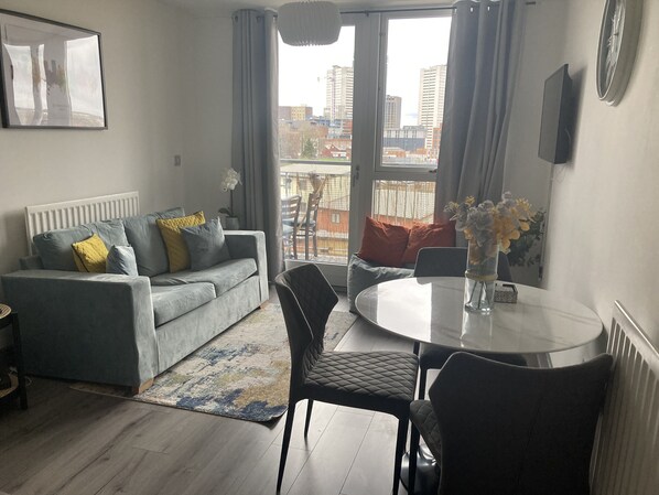 A modern living room with a grey sofa, a round dining table, and a view of the city through large windows. - Lovely City Break at the Lux City 1bed Apartment (Birmingham)