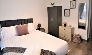 Een keurig opgemaakt slaapkamer met een groot bed, een garderobe, een nachtkastje met een lamp en een klein, rond poefje.
