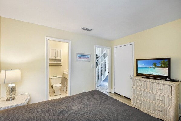 A bedroom with a bed, a TV on a dresser, a bathroom with a toilet and sink, and a staircase leading to another room.