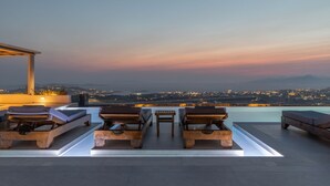 A rooftop lounge with wooden loungers and a pool, overlooking a cityscape at dusk. - Sunset Hill Suites (Mykonos)