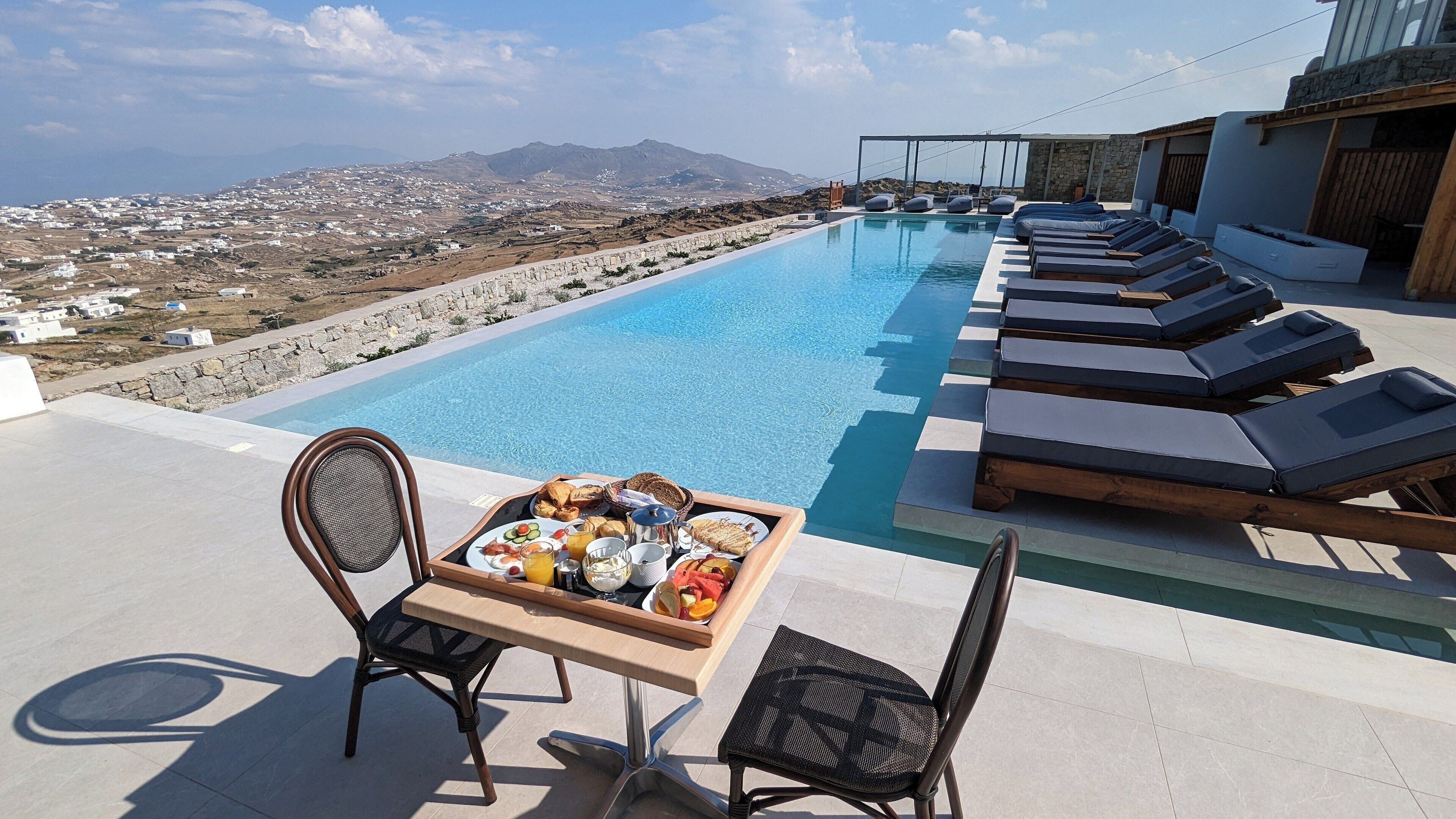 A poolside dining area with a table set for breakfast, overlooking a mountainous landscape.