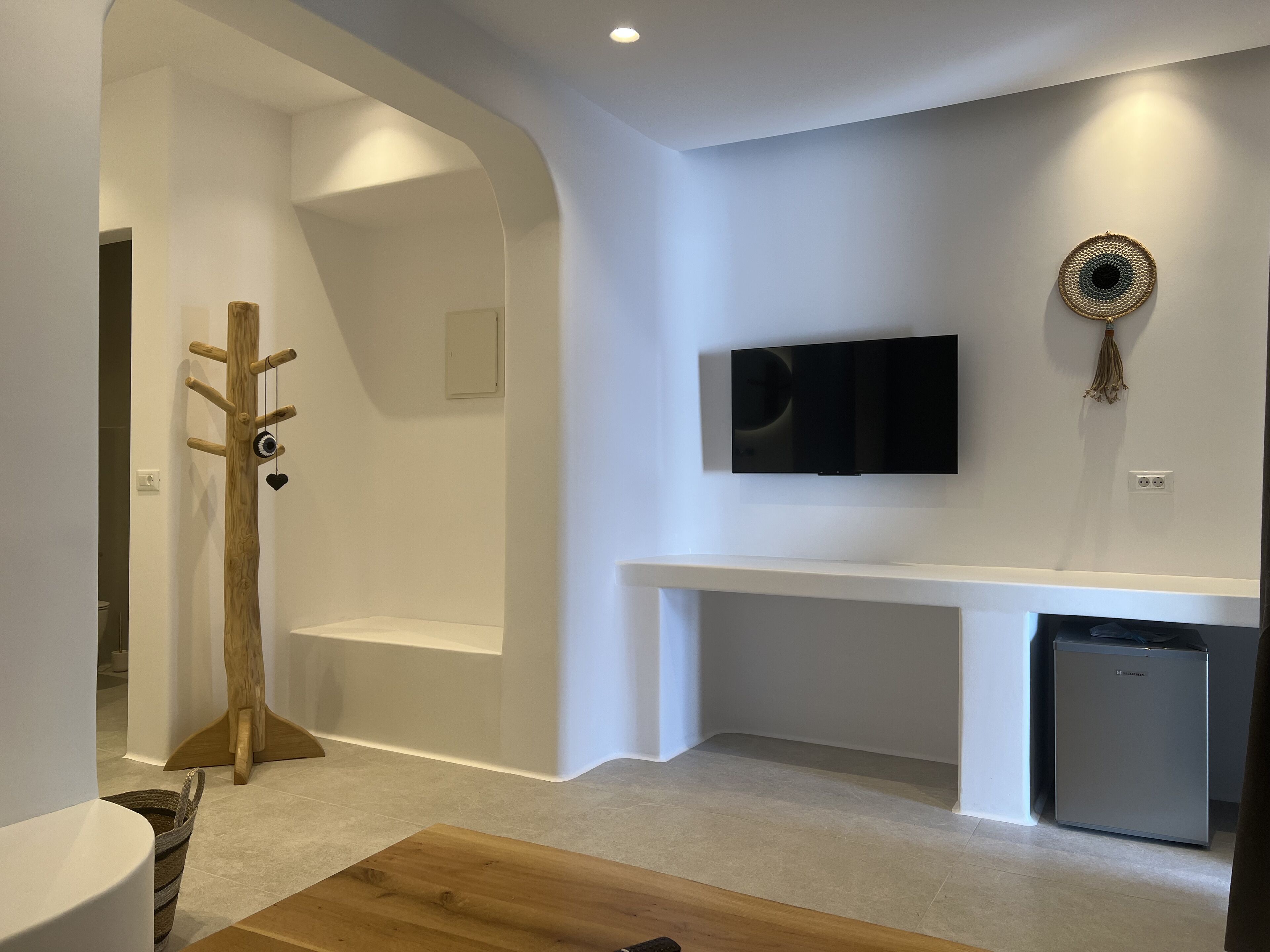 Ein modernes Wohnzimmer mit einem Holzkleiderhaken, einem flachbildfernseher an der Wand, einem kleinen Kühlschrank und einem Holztisch.