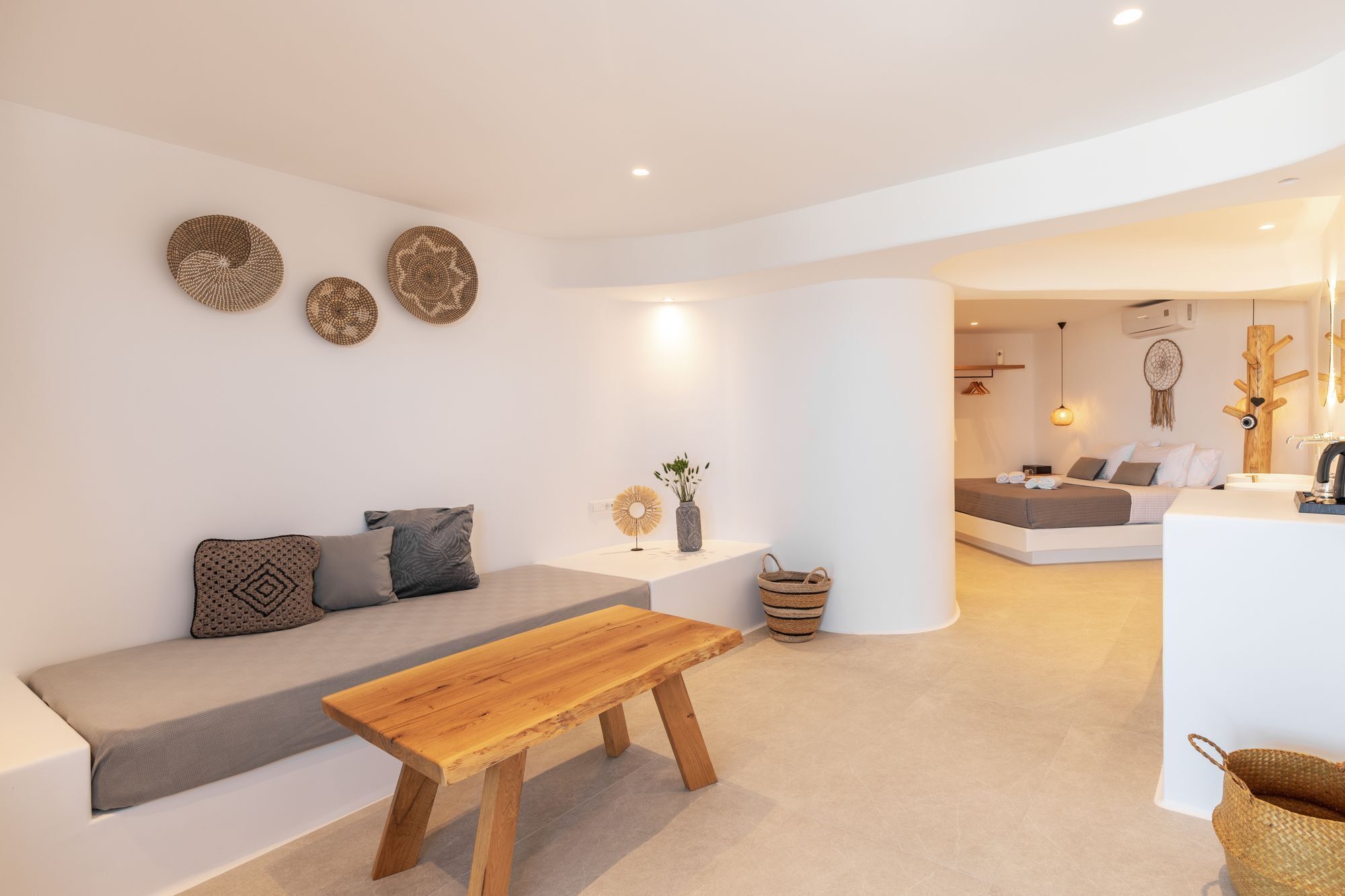 A modern living room with a wooden bench, a grey sofa, and a white coffee table. There are decorative woven baskets and a dreamcatcher on the wall.