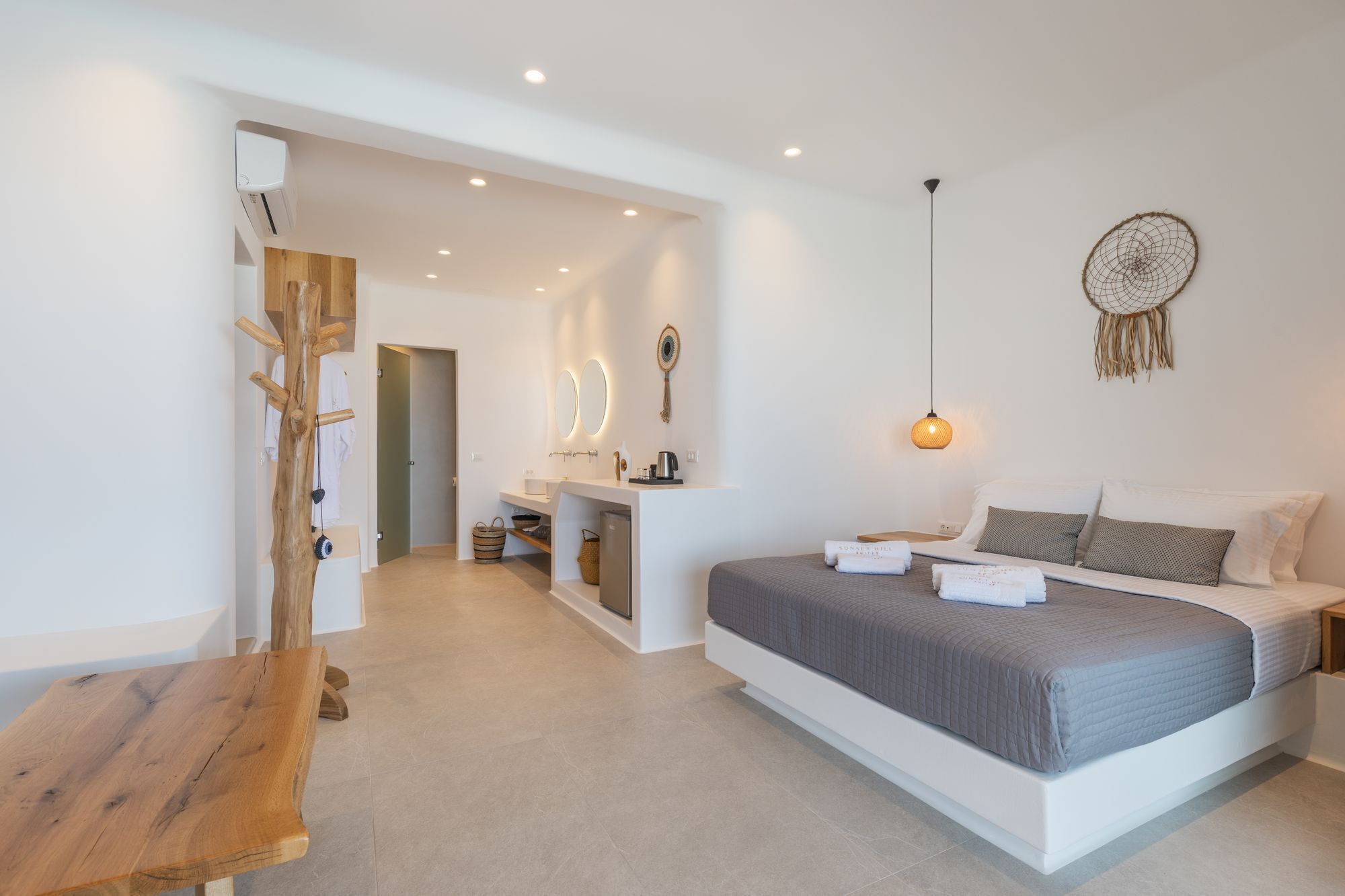 Un dormitorio moderno con una cama grande, un banco de madera, un tocador con espejo y un atrapasueños en la pared.