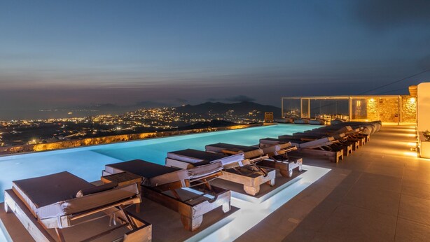 A rooftop infinity pool with lounge chairs and a stone wall. - Sunset Hill Suites (Mykonos)