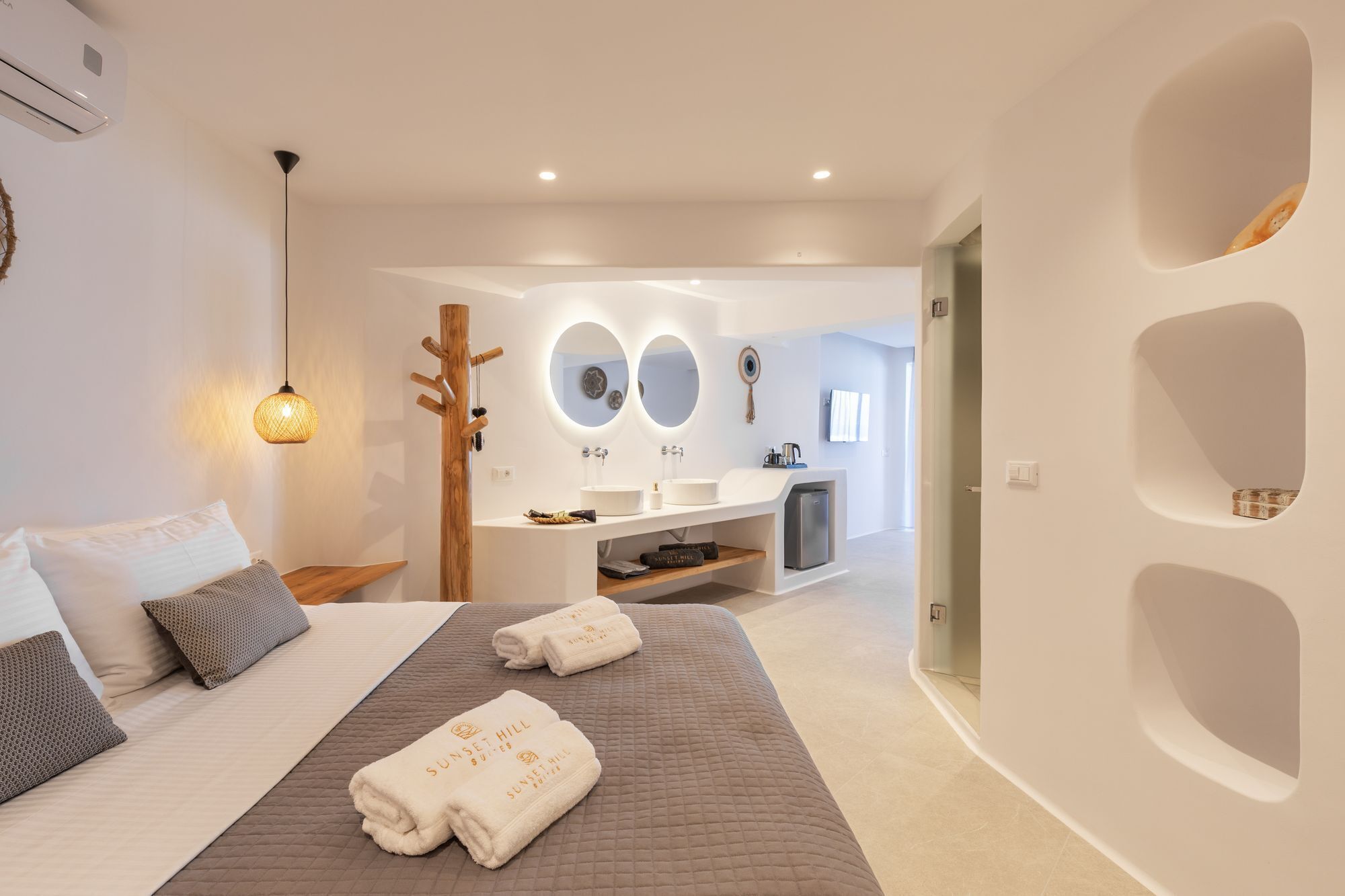 A modern hotel room with a large bed, a wooden coat rack, a bathroom with a sink and mirror, and a unique wall design.