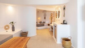 A modern, well-lit room with a wooden table, a white sofa, and a bed with a white headboard. There are wall-mounted lights and a dreamcatcher on the wall. - Sunset Hill Suites (Mykonos)