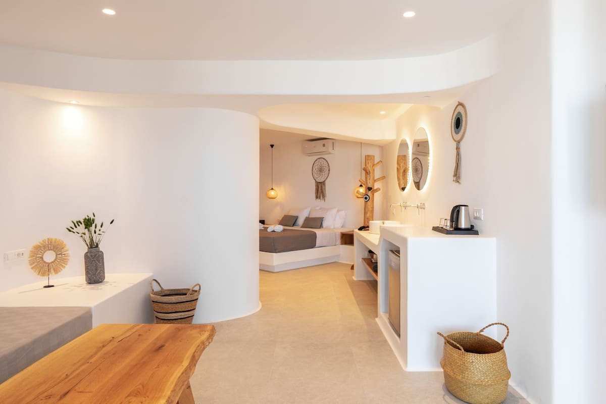 A modern, well-lit room with a wooden table, a white sofa, and a bed with a white headboard. There are wall-mounted lights and a dreamcatcher on the wall.