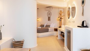 A modern, well-lit room with a wooden table, a bed with a headboard, a built-in bench, and a small kitchen area with a sink and a coffee machine. - Sunset Hill Suites (Mykonos)