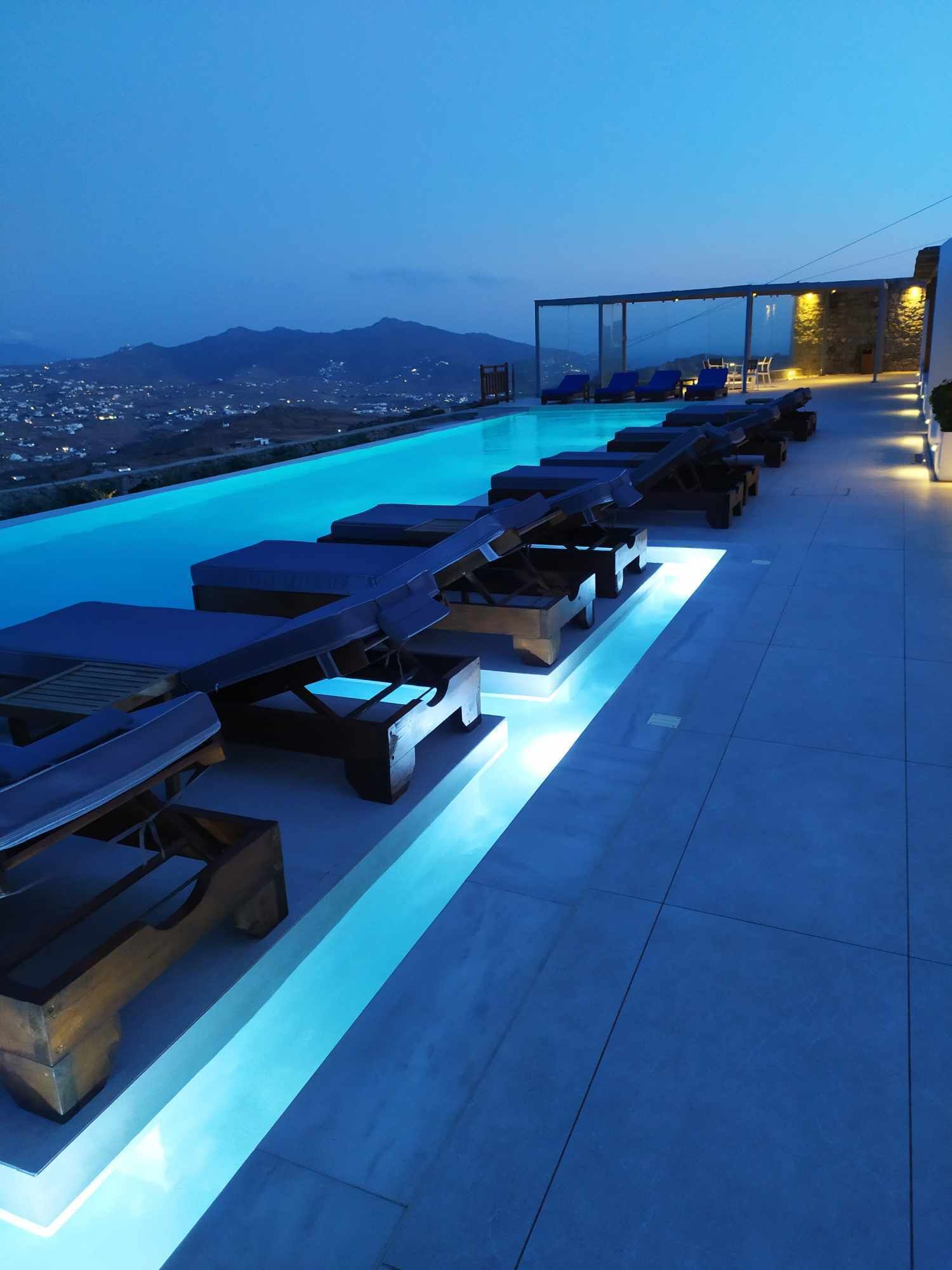 A rooftop infinity pool with lounge chairs and a view of the city and mountains at dusk.