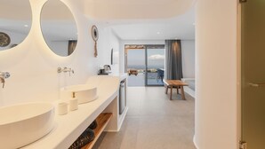 A modern bathroom with a large mirror, two sinks, and a view of a balcony with a table and chairs. - Sunset Hill Suites (Mykonos)
