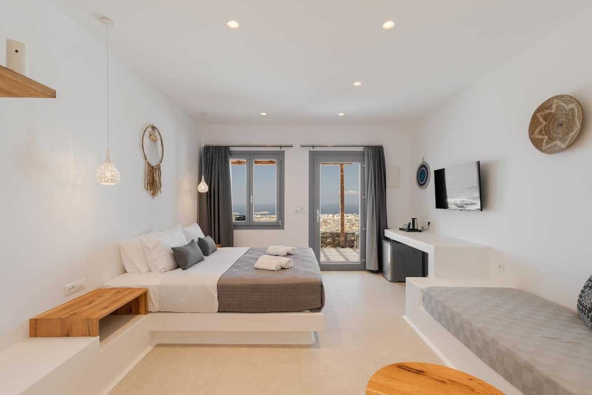 A modern bedroom with a large bed, a smaller bed, a desk, and a TV. There are two windows with curtains, a wall-mounted TV, and a decorative wall hanging.
