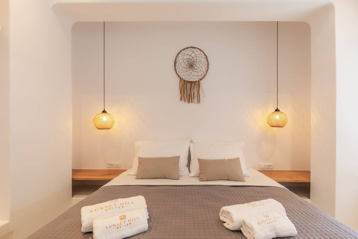 A hotel room with a bed, two bedside tables, a wall-mounted dreamcatcher, and two hanging pendant lights.