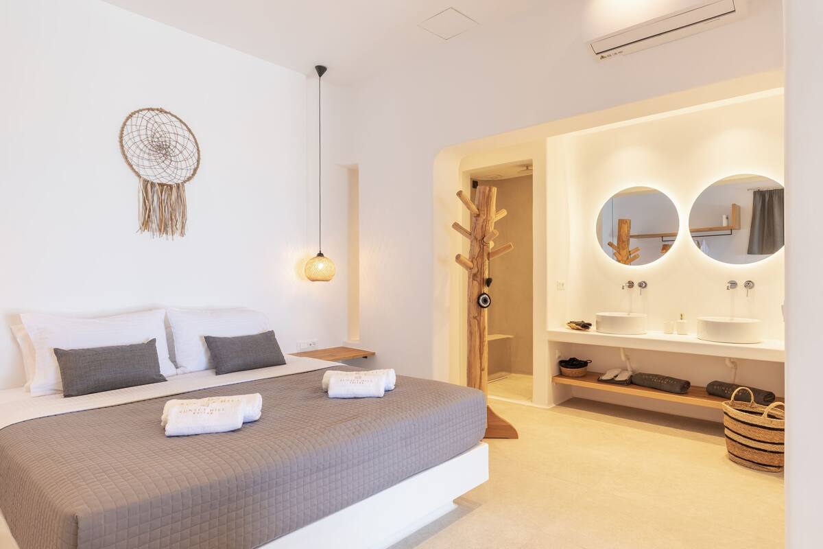 A modern bedroom with a large bed, a bedside lamp, a dreamcatcher, and a bathroom with a vanity and mirrors.
