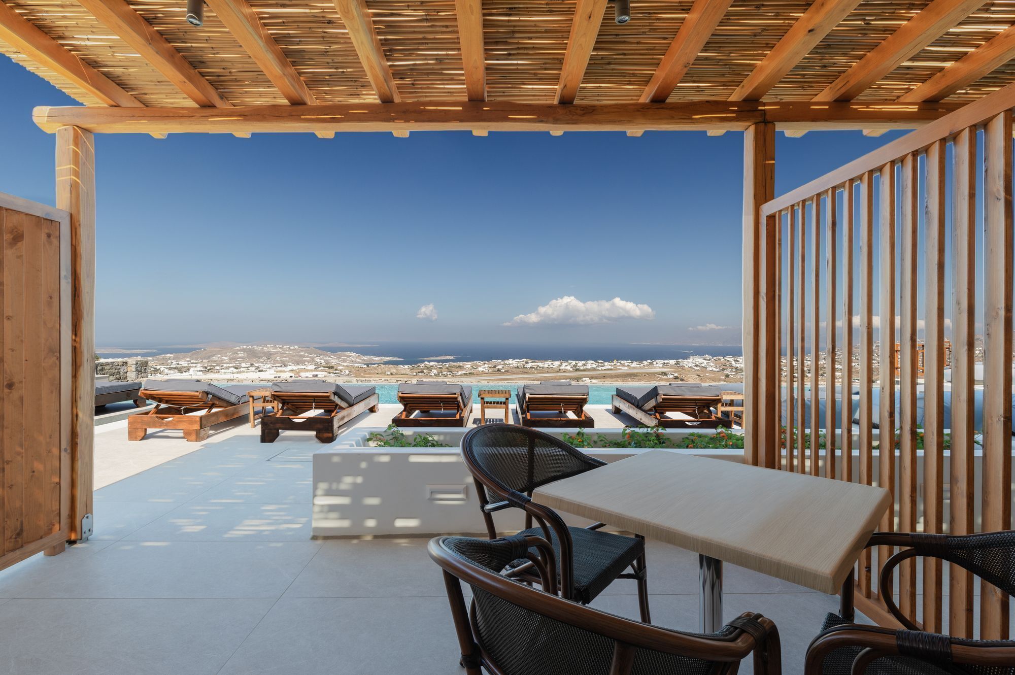 A terrace with wooden furniture, a table, and chairs, overlooking a coastal landscape.