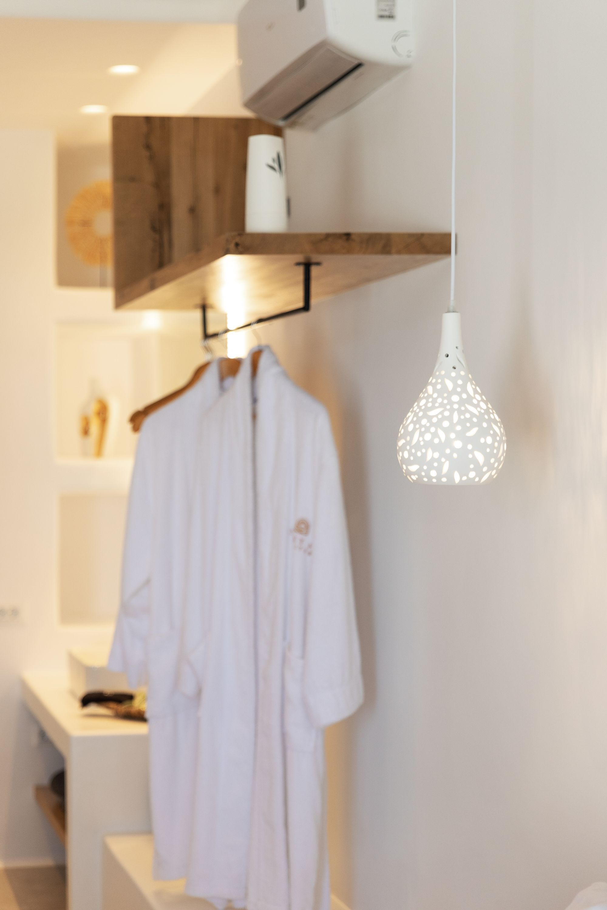 A modern bathroom with a white robe hanging on a rack, a wooden shelf above the sink, and a unique patterned pendant light.