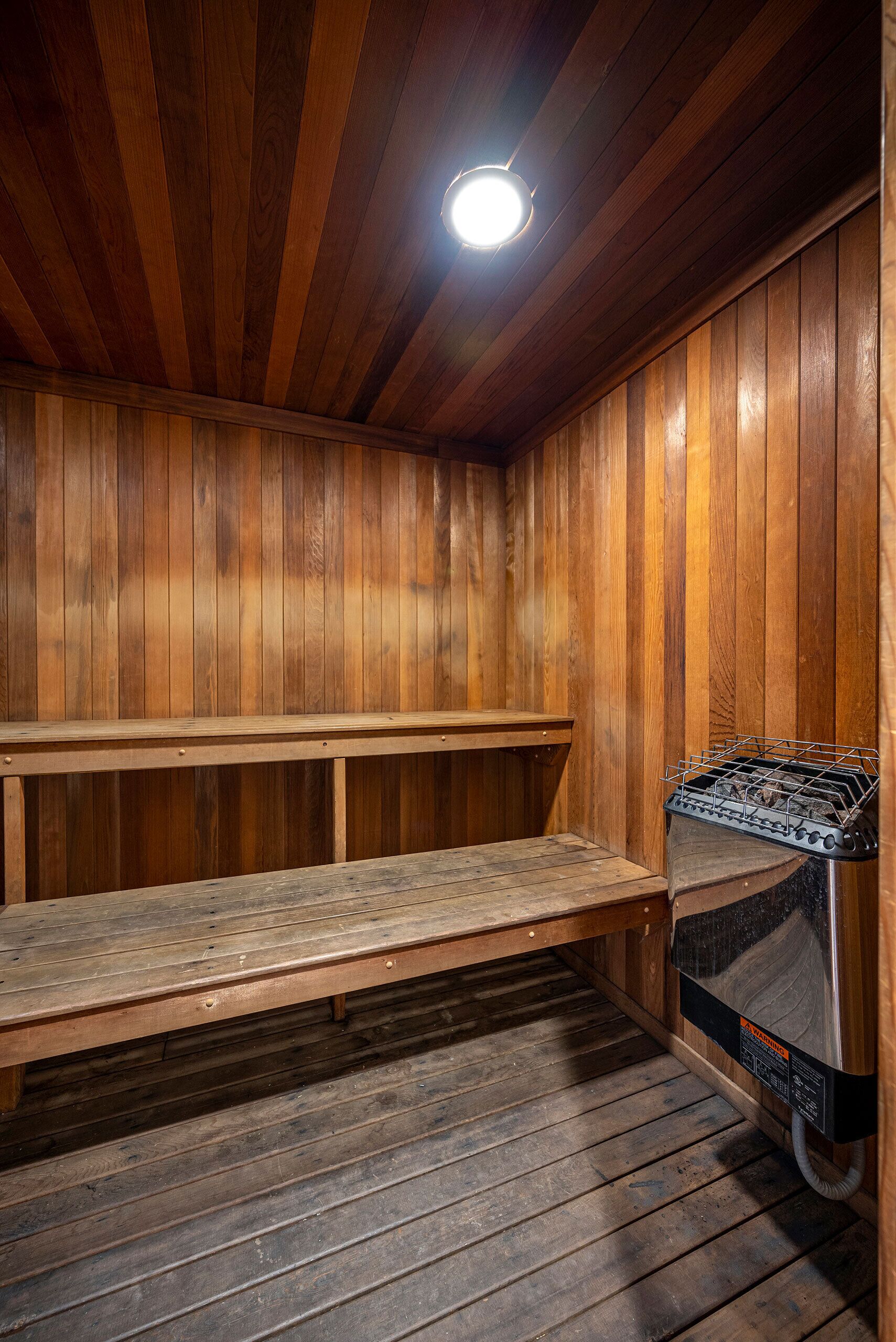 A sauna room with wooden walls and benches, a sauna unit, and a ceiling light.