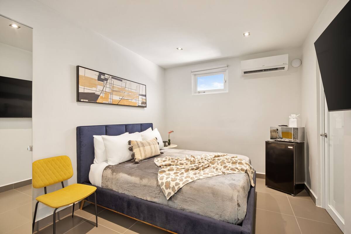 A modern bedroom with a bed, a chair, a small table, an air conditioner, and a window.