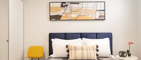 Un dormitorio con una cama, una silla, una mesita de noche con una planta y una obra de arte en la pared.