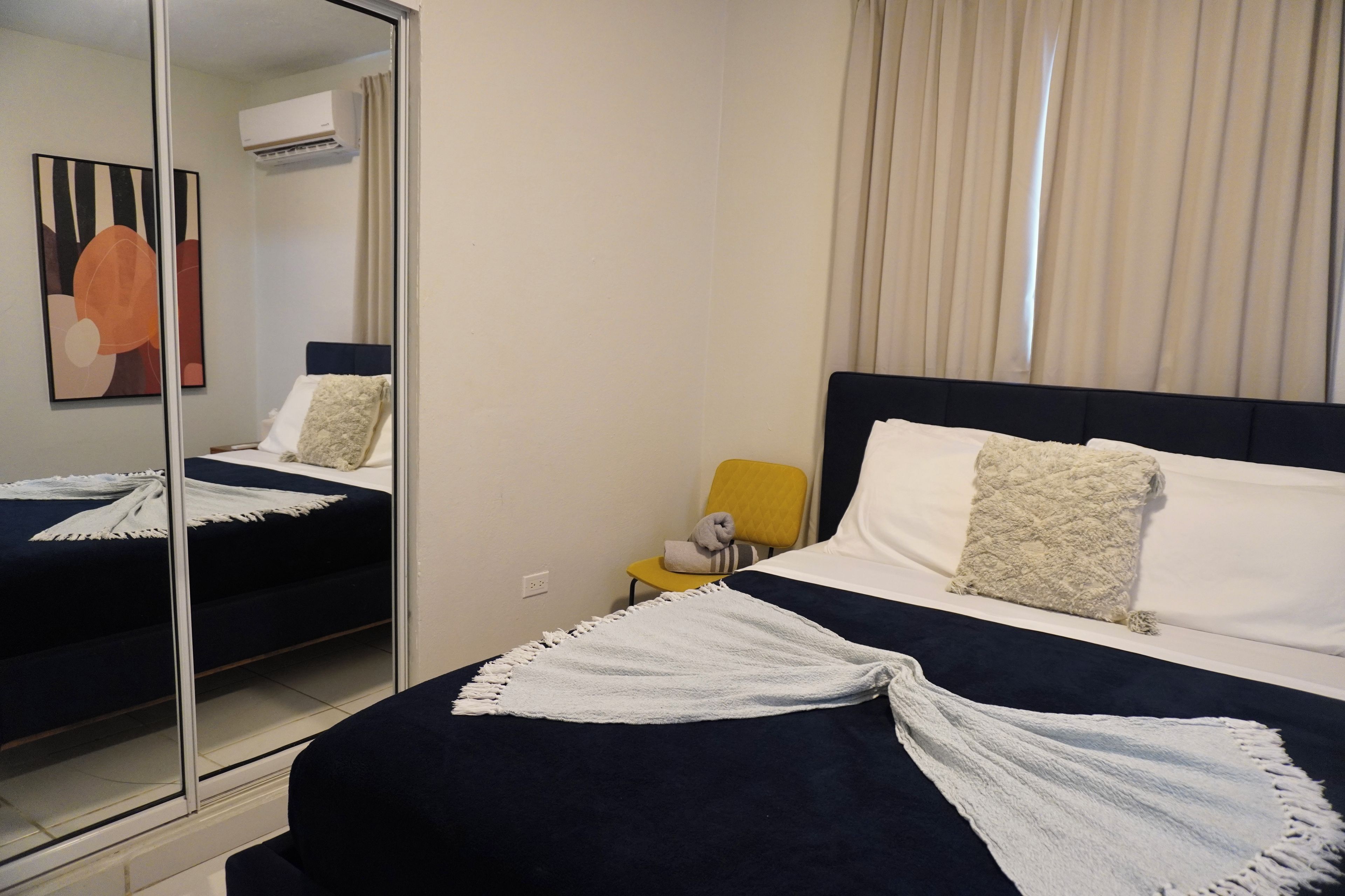 A hotel room with a bed, a mirror, a window with curtains, and a small table with a chair.