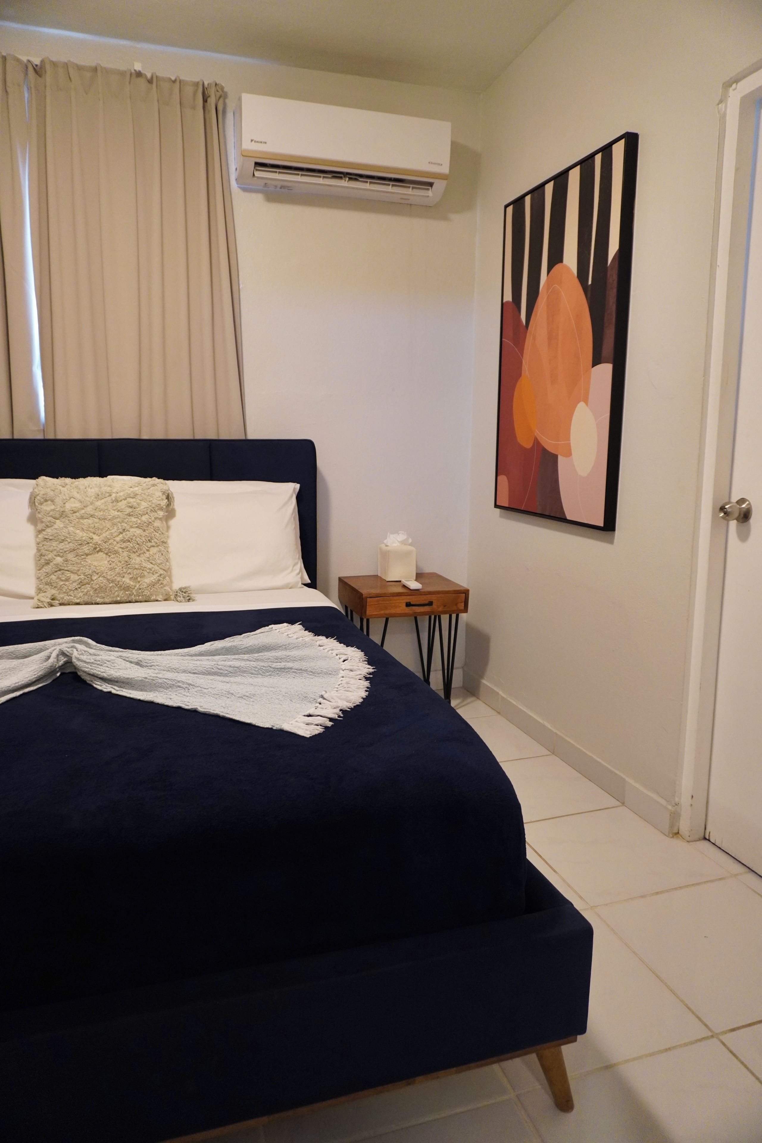 Un dormitorio con cama, aire acondicionado, una mesita con caja de pañuelos y un cuadro en la pared.