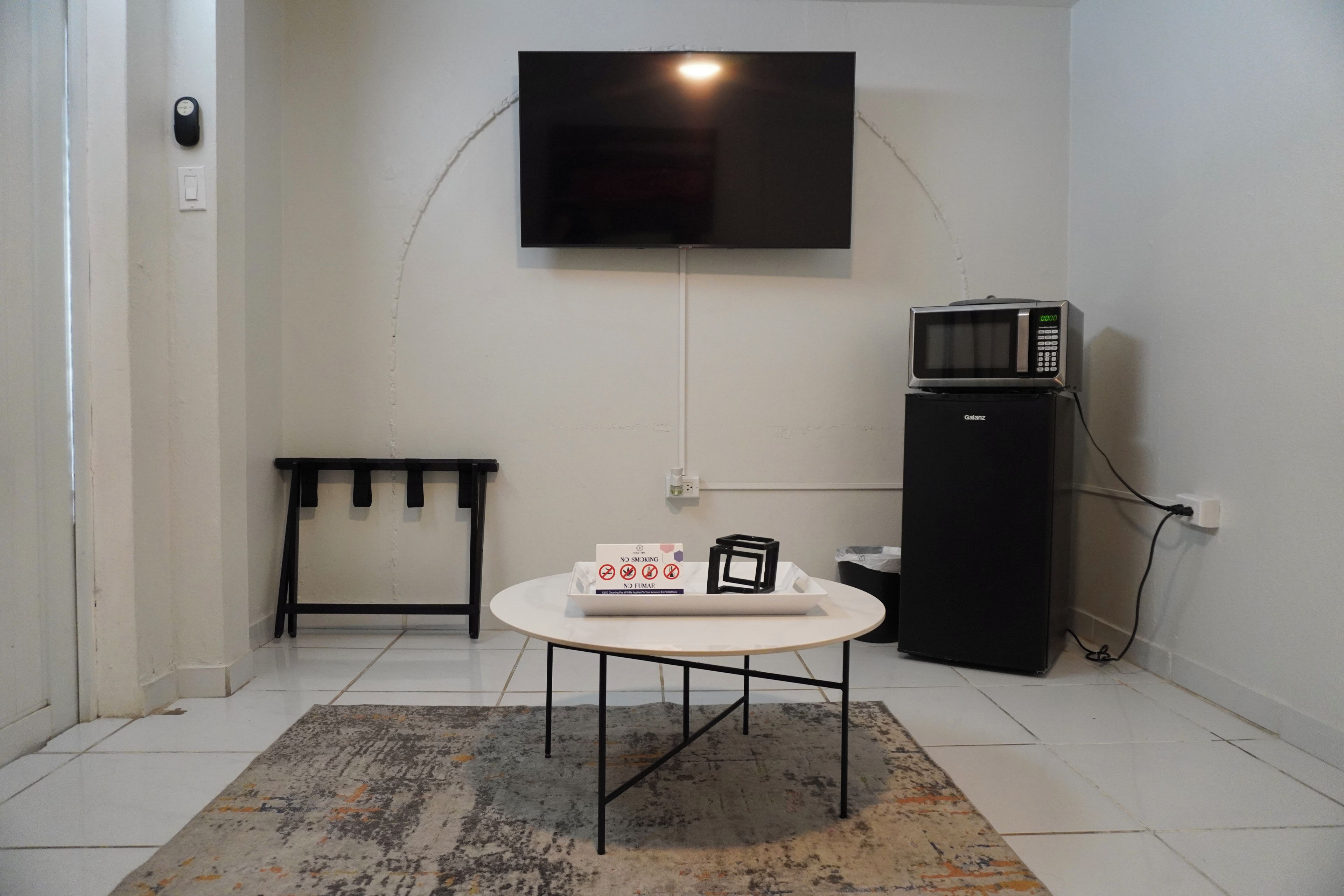 Una habitación con una televisión de pantalla plana montada en la pared, una mesita con una bandeja de objetos, un mueble negro con microondas y un banco contra la pared.