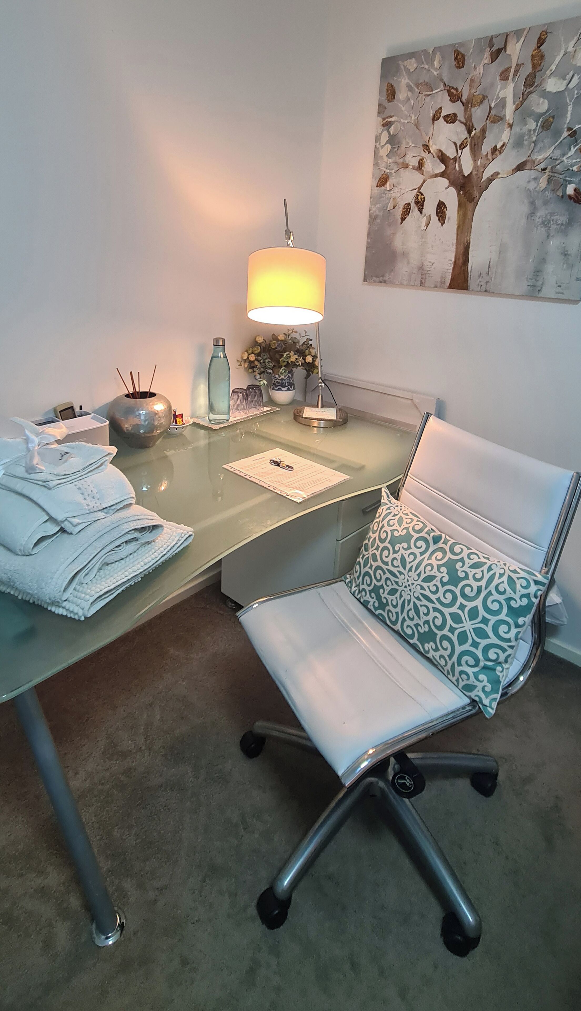 A modern office space with a glass desk, a white office chair with a patterned cushion, a lamp, and a decorative vase with flowers.