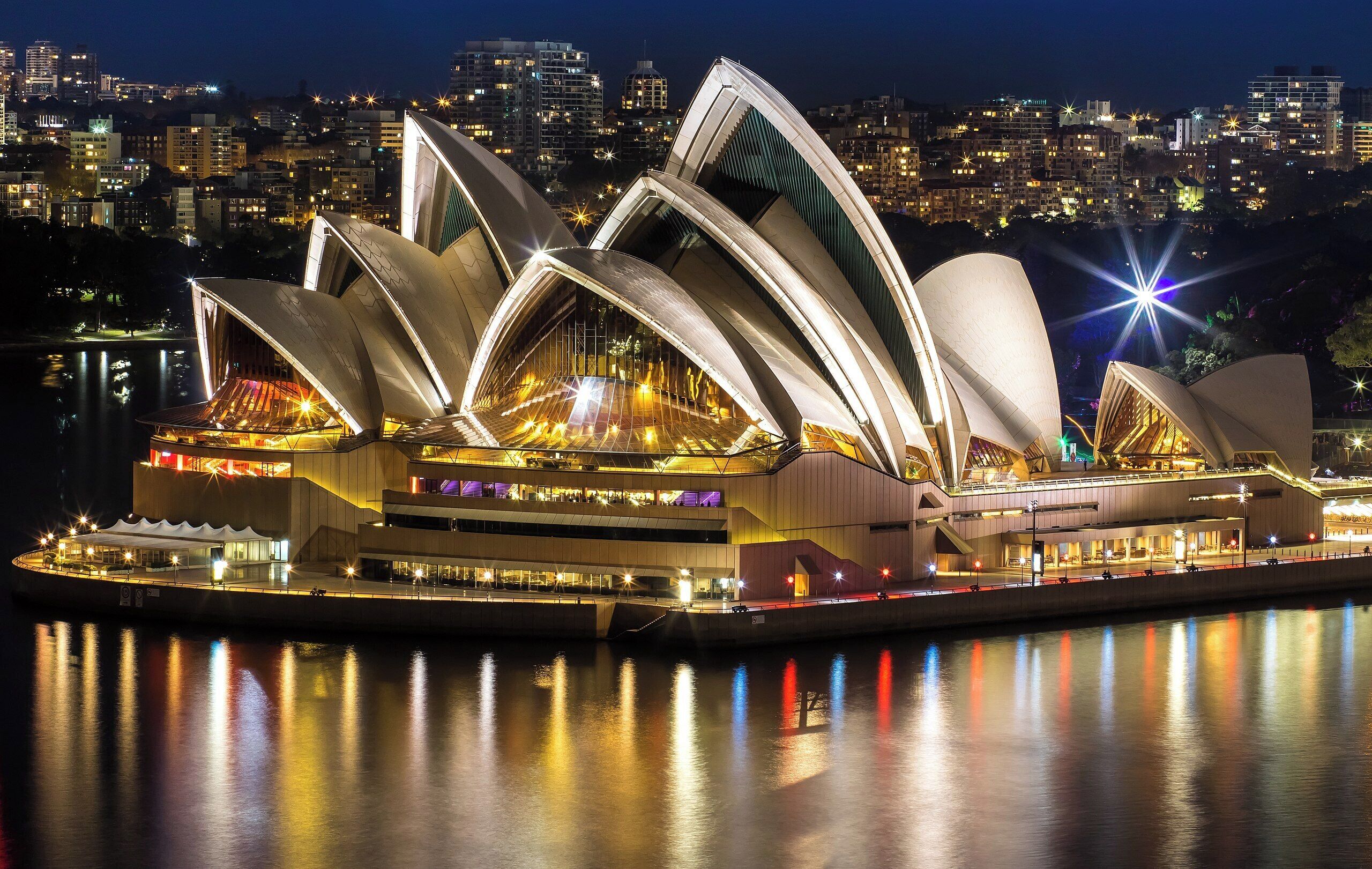 A waterfront building with multiple arched structures and a central stage area, surrounded by water reflecting lights.