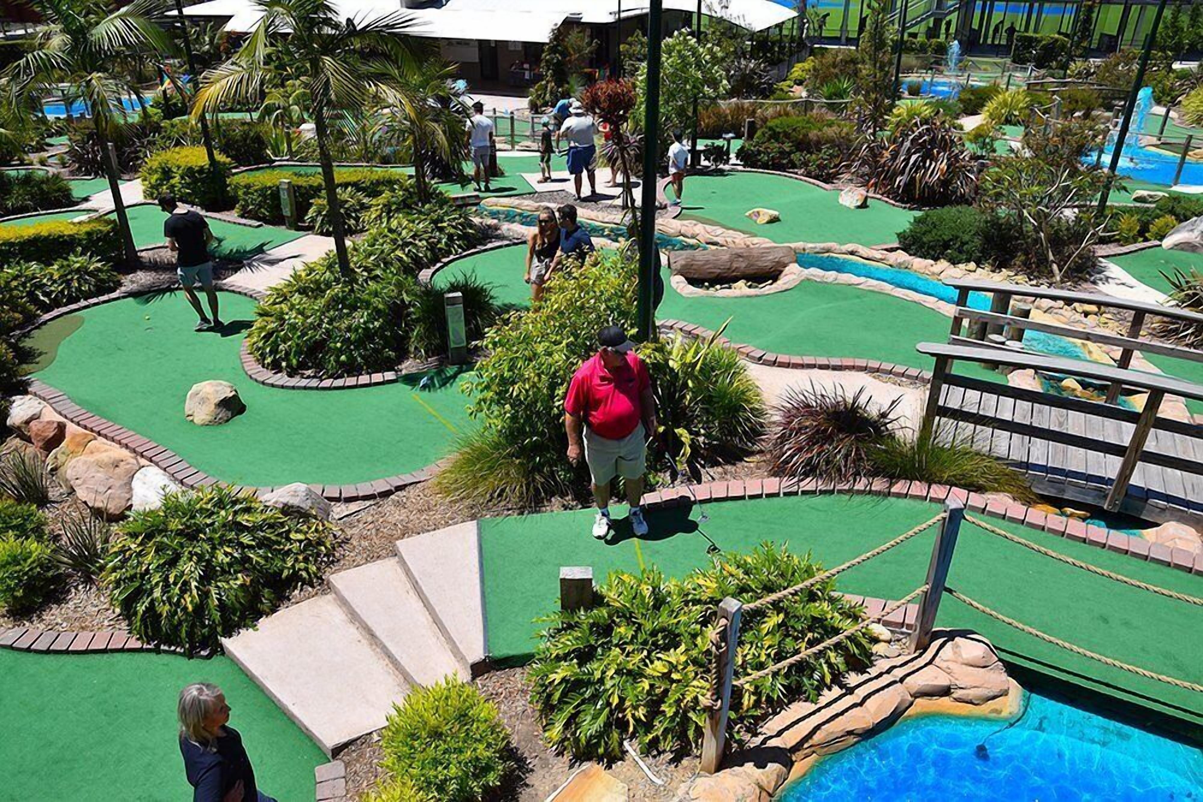 A miniature golf course with a pool, surrounded by tropical plants and a wooden bridge.