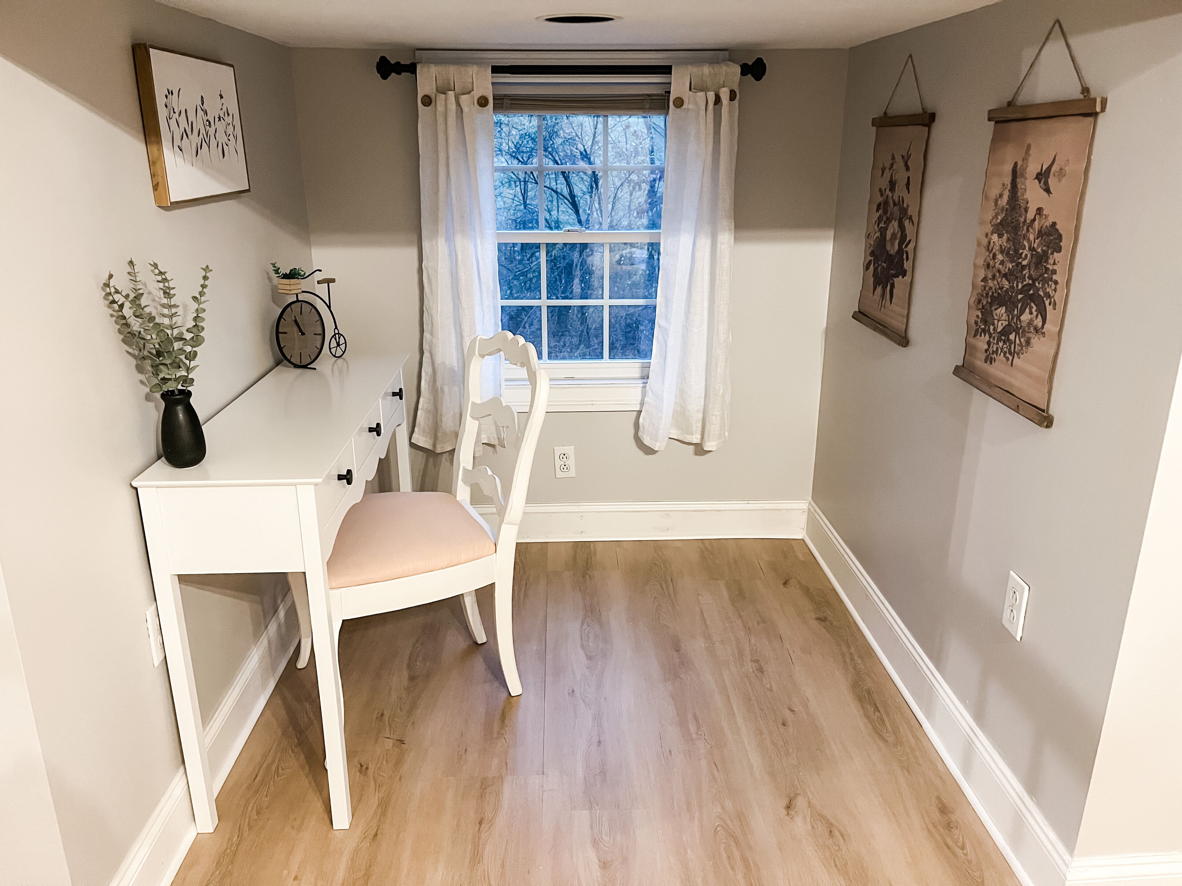 A room with a white desk, a chair, a window with white curtains, and wall-mounted artwork.