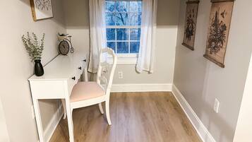 A room with a white desk, a chair, a window with white curtains, and wall-mounted artwork.