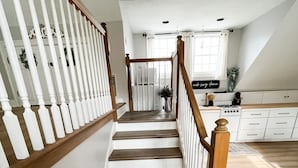 Una escalera con balaustres de madera y barandilla blanca que lleva a una zona de cocina con armarios blancos y un ventanal.