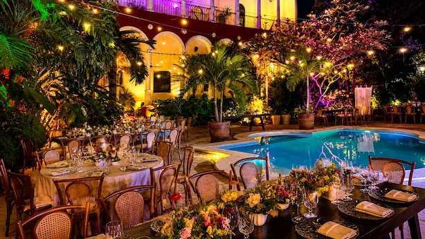 Un espacio exterior con mesas dispuestas para una comida, una piscina y un edificio con arcos al fondo.