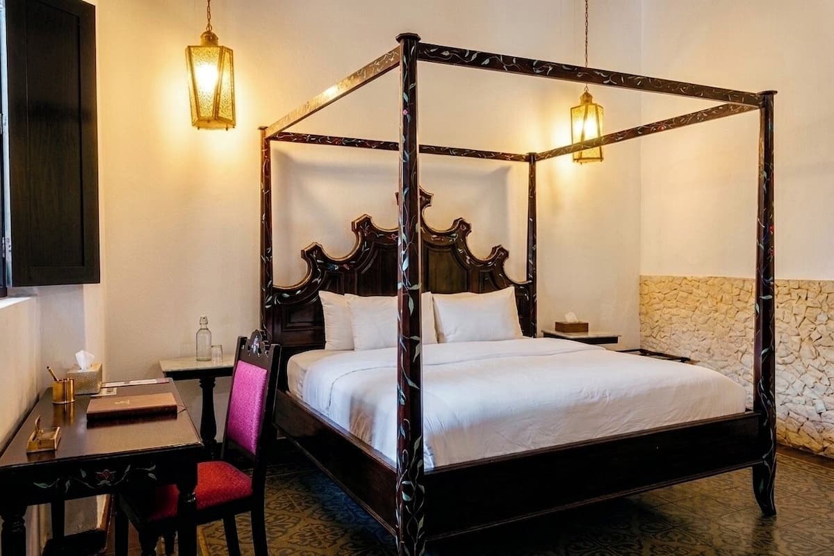 A four-poster bed with a dark wooden frame and white bedding, a small bedside table with a lamp, and a chair with a red cushion.